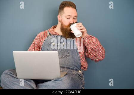 Portrait of brutal bearded hipster man in blue overalls, checked shirt, drinking coffee from a paper cup working on laptop sitting near the wall. Dist Stock Photo