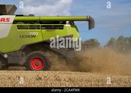 Winter Harvest Close view of rear of combined harvester at work in field Dusty Sunny cloudy sky Fields Trees and houses in distance Landscape format Stock Photo