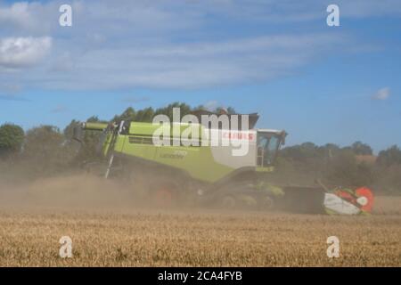 Winter Harvest combined harvester at work in field Dusty Sunny cloudy sky Fields Trees and houses in distance Landscape format Stock Photo