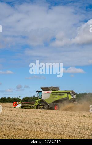 Winter Harvest combined harvester at work in field Dusty Sunny cloudy sky Fields Trees and houses in distance Portrait format Stock Photo