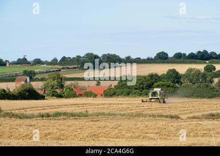 Winter Harvest combined harvester at work in field Dusty Sunny cloudy sky Fields Trees and houses in distance Landscape format Stock Photo