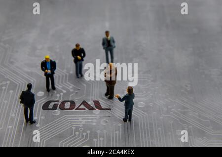 Miniature figurines posed as business people in a meeting over abstract futuristic circuit board with text Goal