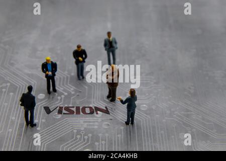 Miniature figurines posed as business people in a meeting over abstract futuristic circuit board with text Vision