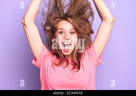 Close up portrait shot of playful young female jumping high, shouting loudly, messy hair upwards, suprised and happy to get present, dreams come true, Stock Photo