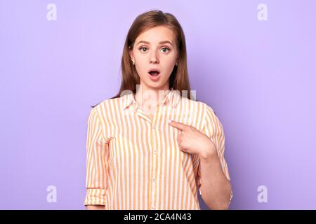 Astonished female teenager points at herself, eyes and mouth wide opened, stupefied face expression, feels unsure and insecure, dressed in comfortable Stock Photo