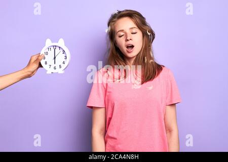 Studio shot of young good looking female in pink pyjamas yawning, trying to wake up, hates early mornings, had bad sleep, disturbing dreams, posing ov Stock Photo