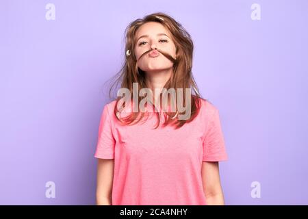 Portrait shot of cheerful funny girl having playful mood, making moustache with her hair, joking and having great time with friends at pyjamas party, Stock Photo