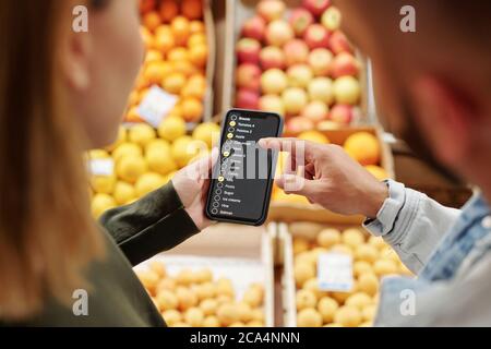 Over shoulder view of young couple checking list of products on smartphone while purchasing food at farmers market Stock Photo
