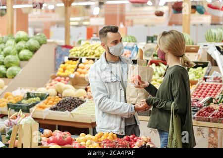 Young couple in cloth masks standing at counter and buying organic food at market: girl choosing tomatoes Stock Photo