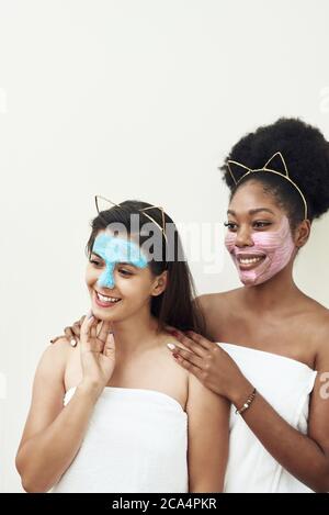 Multimasking. Two different ethnic girls are hugging. The girls' faces are covered with pink and blue face care masks. Beauty face, facial treatment and spa concept. Stock Photo