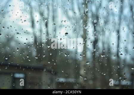 Raindrops on the window glass on a gloomy day. In the background, blurred silhouettes of trees and houses. Autumn. Stock Photo