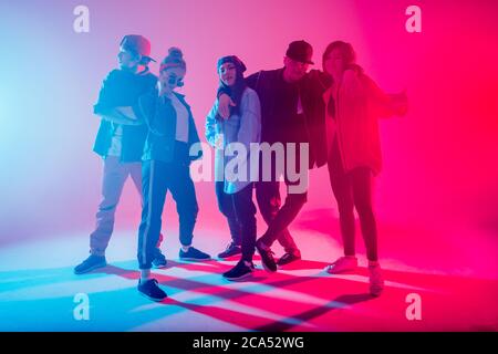 Young modern dancing group of five adult young people practice dancing on colorful background. Fashionably dressed youngsters posing over blurred disc Stock Photo