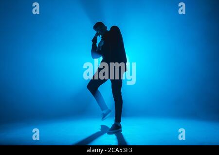 Silhouette of man giving solo performance, dancing alone in hip hop style on club scene with blue neon lightning and smoke. Stock Photo