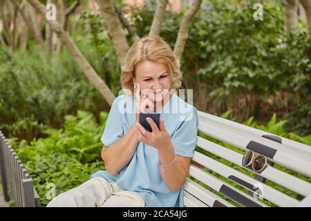 Aged caucasian attractive woman about 60 years old is sitting on the bench outdoor and looking in her smartphone. She is smiling and looks happy and c Stock Photo