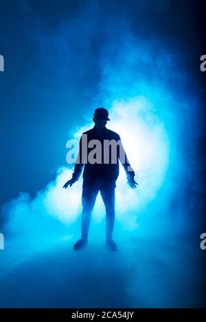Silhouette of man giving solo performance, dancing alone in hip hop style on club scene with blue neon lightning and smoke. Stock Photo