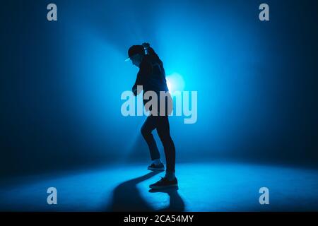 Silhouette of man giving solo performance, dancing alone in hip hop style on club scene with blue neon lightning and smoke. Stock Photo