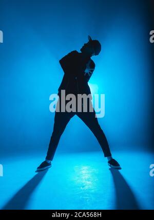 Silhouette of man giving solo performance, dancing alone in hip hop style on club scene with blue neon lightning and smoke. Stock Photo