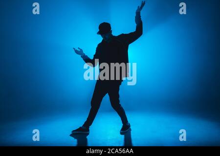 Silhouette of man giving solo performance, dancing alone in hip hop style on club scene with blue neon lightning and smoke. Stock Photo