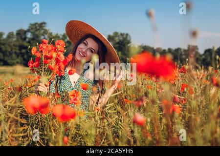 Young smiling woman picking bouquet of poppies flowers walking in summer field. Happy girl feeling free