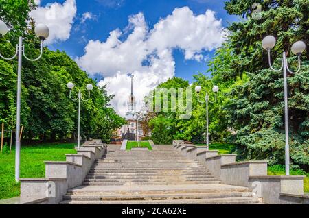 Staircase in green trees alley with street lights, police men monument and House under the Spire in Minsk city historical centre, blue sky white clouds in sunny summer day, Republic of Belarus Stock Photo