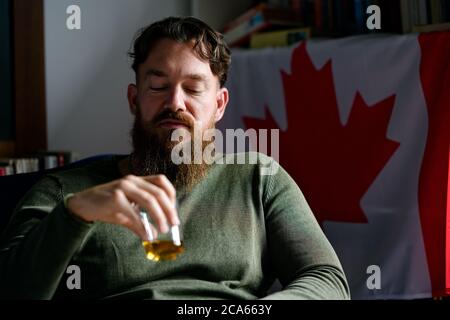 Dark tonal close up portrait of a young stylish handsome bearded Canadian man enjoying whiskey in a room with a Canadian flag behind him Stock Photo