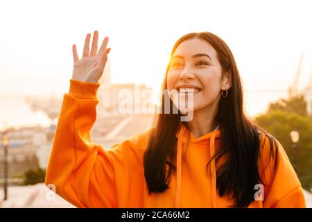 Photo of joyful beautiful asian woman smiling and waving hand while walking outdoors Stock Photo