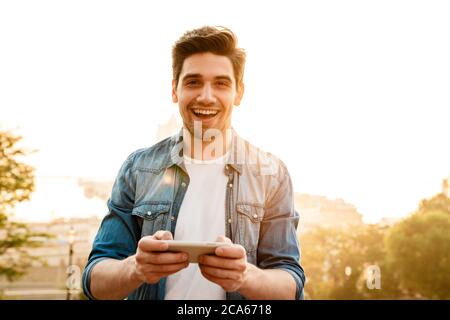 Photo of excited handsome man smiling and playing online game on mobile phone while walking outdoors Stock Photo