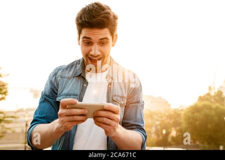 Photo of excited handsome man smiling and playing online game on mobile phone while walking outdoors Stock Photo