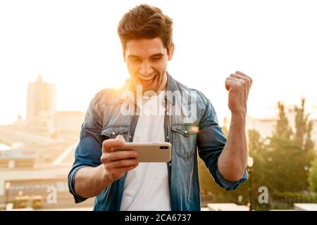 Photo of excited man making winner gesture and playing online game cellphone while walking outdoors Stock Photo