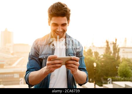 Photo of excited handsome man smiling and playing online game on mobile phone while walking outdoors Stock Photo