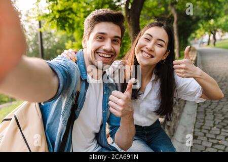 Image of joyful multicultural couple taking selfie photo and showing thumbs while sitting on bench in park Stock Photo