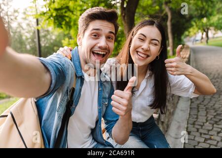 Image of joyful multicultural couple taking selfie photo and showing thumbs while sitting on bench in park Stock Photo