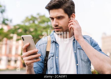 Photo of focused man with wireless earphone using cellphone while walking with backpack on boulevard Stock Photo