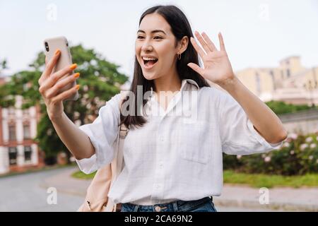 Photo of happy asian woman using cellphone and waving hand while walking on boulevard Stock Photo