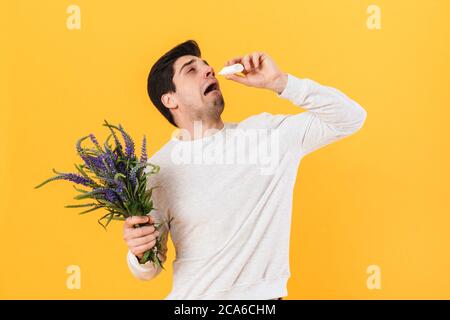 Photo of unhappy man with allergy using nose drops while holding flowers isolated over yellow background Stock Photo