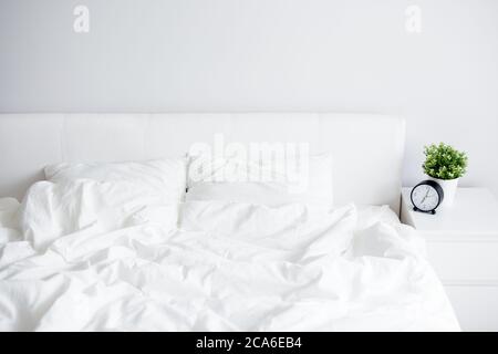 unmade bed with white blanket and two pillows, bedside table with alarm clock and plant pot at home or hotel Stock Photo