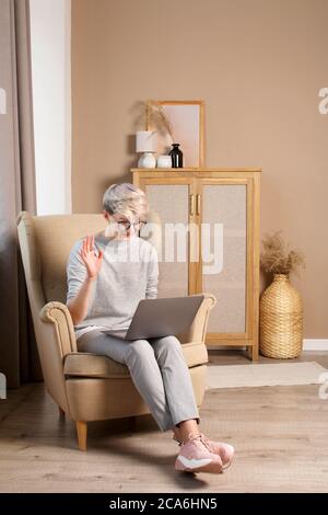 Full body of pretty smart woman about 30s in casual clothes with short blonde hair sitting in a chair and communicating via video call on a laptop Stock Photo