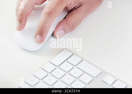 male hand clicking on a white mouse with a white keyboard on the right. White table in a office. Technology and business