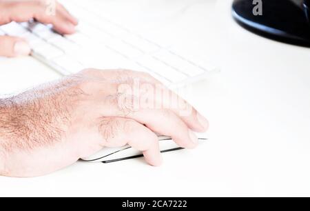 male hand clicking on a white mouse with a white keyboard on the right. White table in a office. Technology and business Stock Photo