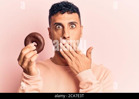 Young hispanic man holding donut covering mouth with hand, shocked and afraid for mistake. surprised expression Stock Photo