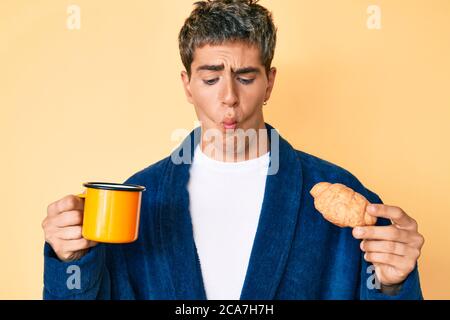 Young handsome man wearing bathrobe eating breakfast holding coffee and croissant making fish face with mouth and squinting eyes, crazy and comical. Stock Photo