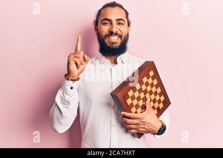 Young arab man holding chess smiling with an idea or question pointing finger with happy face, number one Stock Photo