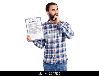 Young arab man holding clipboard with agreement document serious face thinking about question with hand on chin, thoughtful about confusing idea Stock Photo