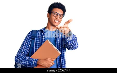 Young african american man wearing student backpack holding book smiling cheerful offering palm hand giving assistance and acceptance. Stock Photo