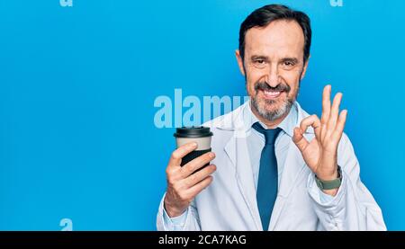 Middle age handsome doctor man wearing coat drinking cup of takeaway coffee doing ok sign with fingers, smiling friendly gesturing excellent symbol Stock Photo