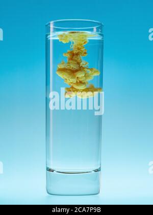 Yellow acrylic paint in water. Abstract ink cloud in glass of water on blue background. Stock Photo