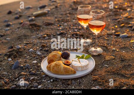 Horizontal image of a romantic beach picnic for two at sunset with two glasses of rose wine, croissants, camamber cheese and figs. Negative space. Stock Photo