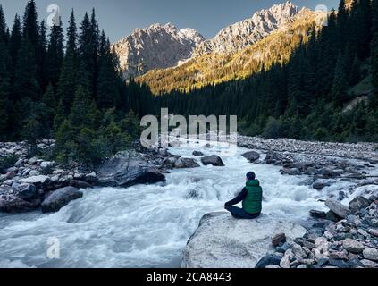 Young man doing yoga meditation in Padmasana sitting position on the rock near the river with mountain at sunrise at background. Stock Photo