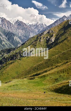 Small tourist walking on green hill in the mountain valley with high snowy peaks Stock Photo