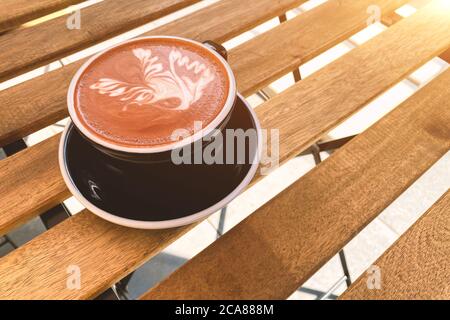 Black cup of cappuccino or cocoa drink with latte art on wooden table, against sunlight. Beautiful latte foam. Place for text Stock Photo